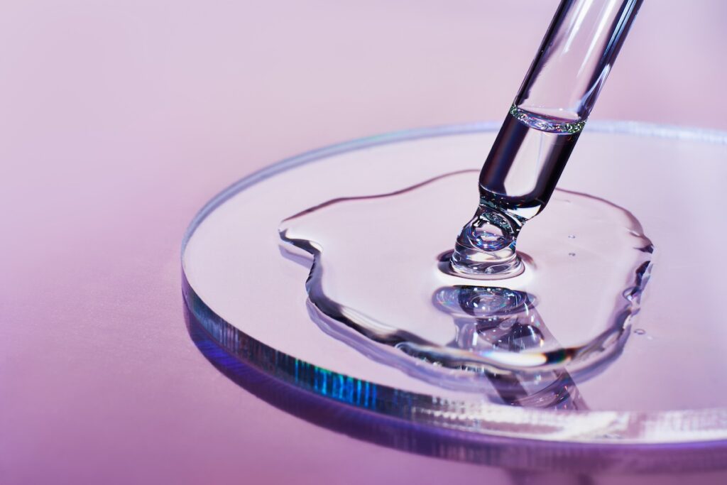 A pipette dips into a pool of clear liquid on a petri dish on a glowing, iridescent field of light.
