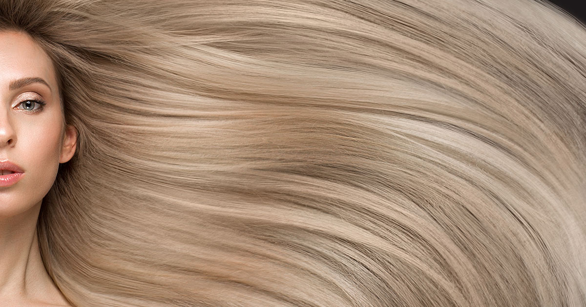 A photo of a woman with incredibly long smooth waves of golden bleach blonde hair. Her face is positioned so it is cut in half by the left edge of the image, with her hair filling all of the image and flowing past the right right. It requires innovation and R&D in beauty contract manufacturing to produce stunning natural results like this.
